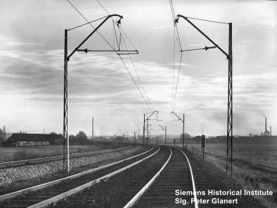 zweigleisige gerade Strecke mit Oberleitung an Streckmetallmasten mit Schrägausleger und einfachen Kappenisolatoren