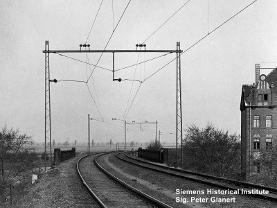 zweigleisige Strecke im Rechtsbogen mit Oberleitung an Stahlmasten und mit Jochtraversen darauf stehenen Dreieraggregaten